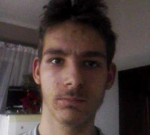 Portret młodego mężczyzny z krótkimi, ciemnymi włosami, patrzącego prosto w obiektyw, na tle jasnego wnętrza z widoczną szafką i firanką.