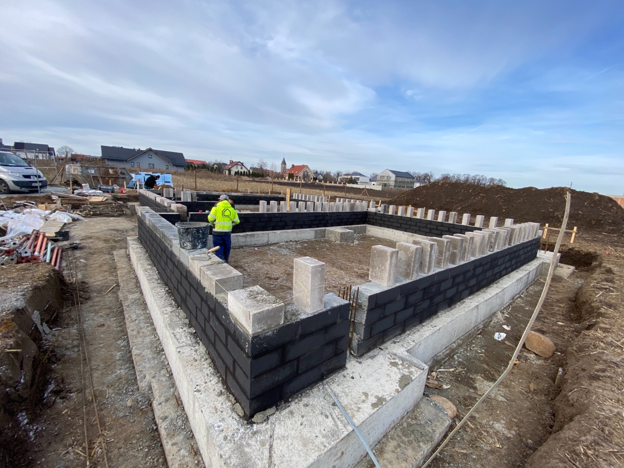 Wznoszenie fundamentów z czarnej cegły na betonowej podstawie, widoczny pracownik w żółtej kamizelce na placu budowy, w tle domy i pochmurne niebo.