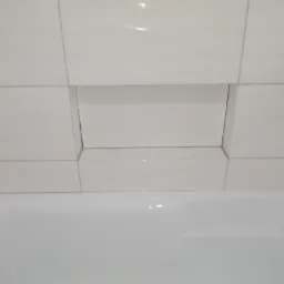 Białe, błyszczące płytki ceramiczne ułożone na ścianie nad wanną, z wnęką wykończoną tymi samymi płytkami, tworzącą półkę.