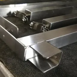 Elementy konstrukcji ze stali nierdzewnej, w tym profile zamknięte i uchwyty zaciskowe, leżące na ciemnej powierzchni roboczej.