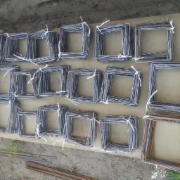 Kwadratowe elementy zbrojeniowe, ułożone w rzędach, związane białym drutem. Widoczne pręty zbrojeniowe w dolnej części kadru, surowy beton w tle.