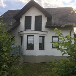 Dwupiętrowy dom z białymi ścianami, ciemną dachówką i szarymi roletami w oknach, otoczony zielenią krzewów i drzew.