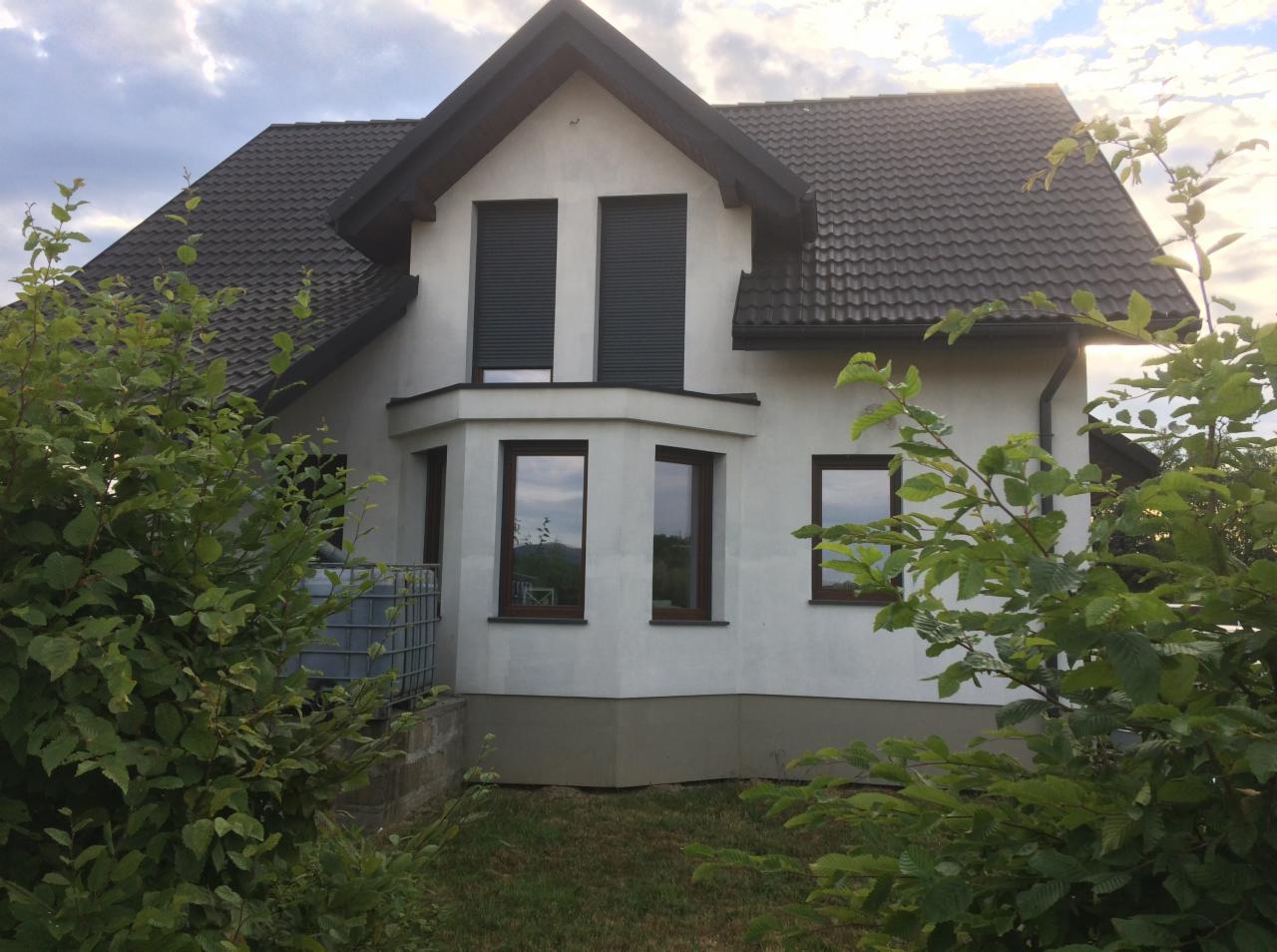 Dwupiętrowy dom z białymi ścianami, ciemną dachówką i szarymi roletami w oknach, otoczony zielenią krzewów i drzew.