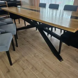 St&oacute;ł z drewnianym blatem i czarną, metalową podstawą w kształcie litery X, otoczony krzesłami z szarą tapicerką, na podłodze panele.