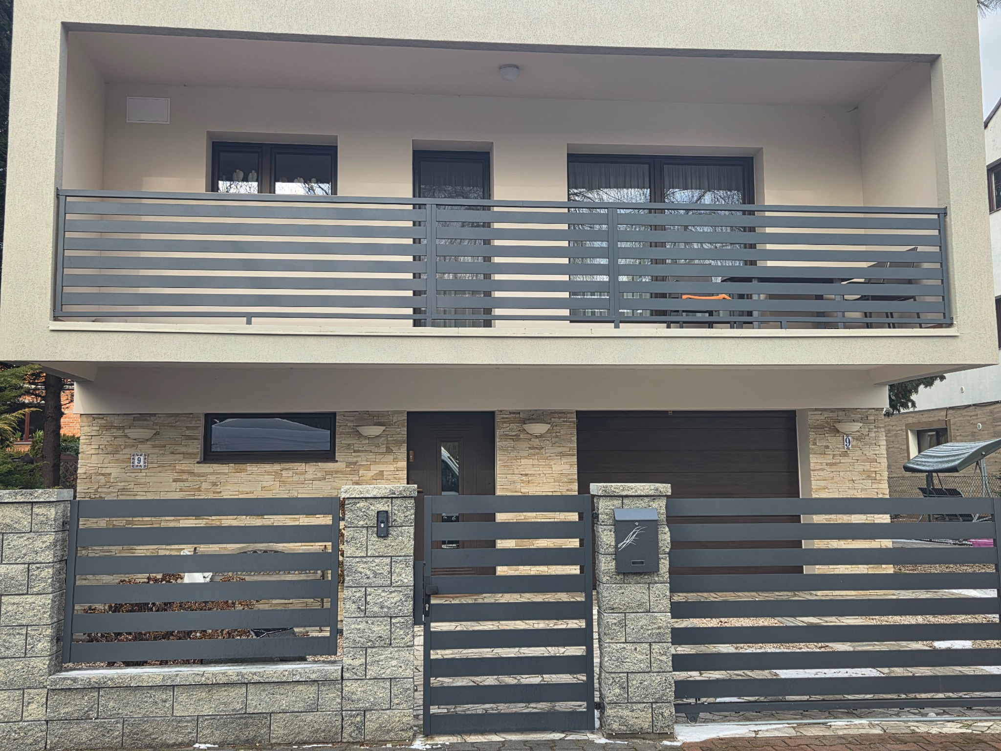 Nowoczesne, poziome, szare ogrodzenie posesji z kamiennymi słupkami i balkon z poziomymi szprosami, na tle dwukondygnacyjnego domu z elewacją w odcieniach beżu i szarości.