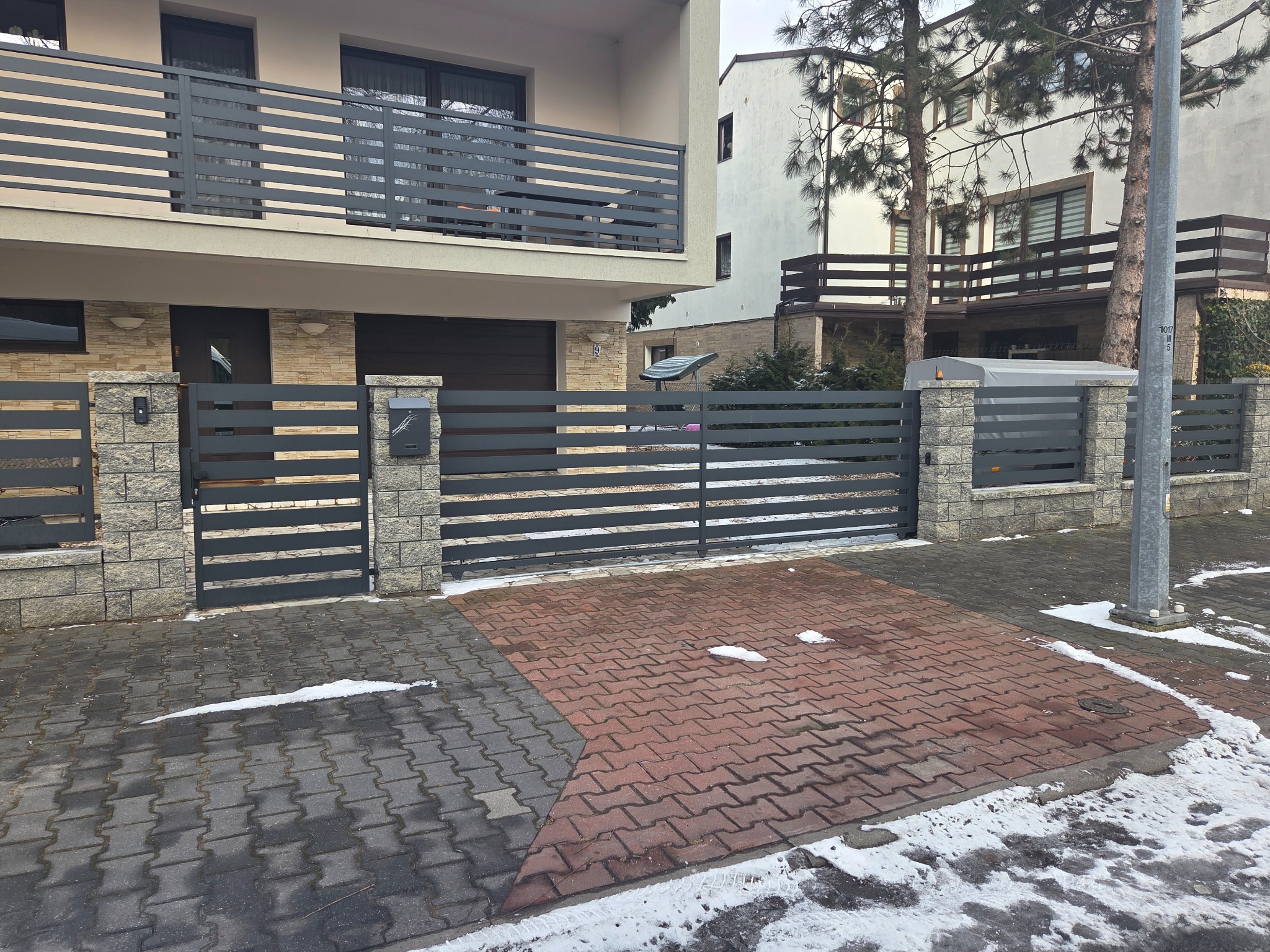 Nowoczesne, poziome, metalowe ogrodzenie posesji z bramą i furtką, osadzone na kamiennych słupkach. W tle dom z balkonem z identycznym ogrodzeniem.