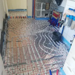 Rozłożona instalacja ogrzewania podłogowego na folii z siatką, widoczne rury doprowadzające wodę i rozdzielacz w tle, wiadro na podłodze.