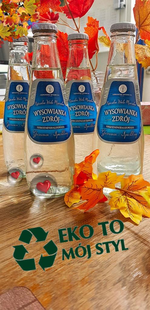 Cztery szklane butelki wody Wysowianka Zdrój z etykietami w odcieniach niebieskiego, ustawione na drewnianej powierzchni z napisem 'Eko to mój styl' i motywem recyklingu, dekorowane jesiennymi liśćmi.