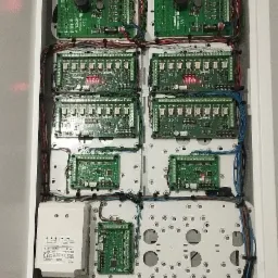 Rozdzielnica z modułami elektronicznymi systemu zabezpieczeń, widoczne liczne kable, płytki drukowane, transformator i akumulatory Alarmtec.