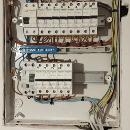 IDSG Adam Witoszek - Otwarta skrzynka elektryczna z zamontowanymi bezpiecznikami Schneider Electric, widoczne kable w kolorowych izolacjach i uszkodzenia tynku wokół skrzynki.