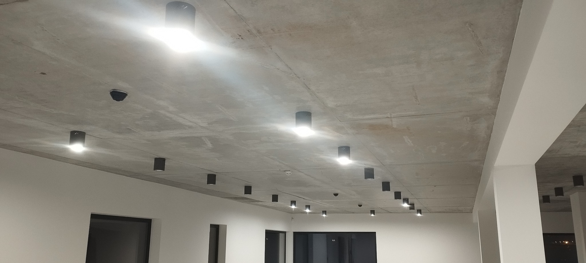 Betonowy sufit z rzędem czarnych, cylindrycznych lamp LED i czujnikiem dymu, oświetlających puste, białe pomieszczenie z ciemnymi otworami okiennymi.