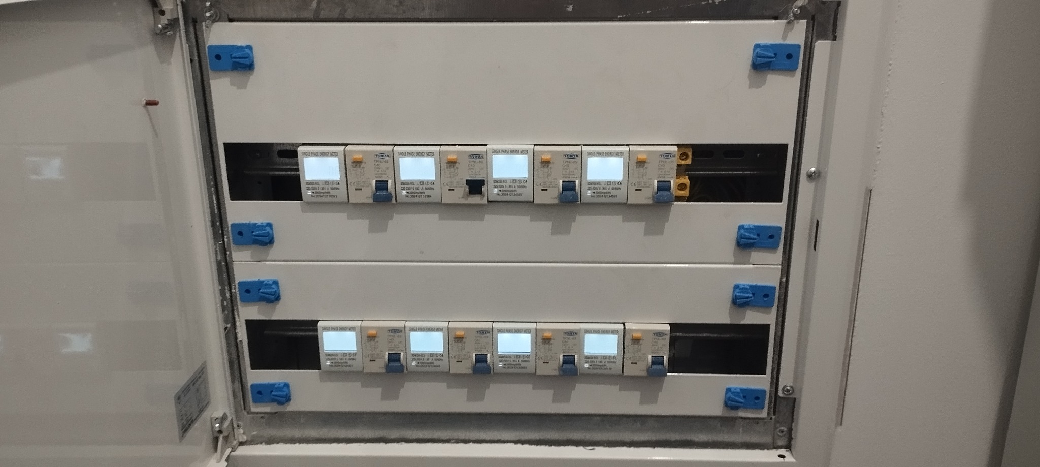 Biała skrzynka rozdzielcza z zamontowanymi licznikami energii elektrycznej, widoczne niebieskie zatrzaski mocujące.