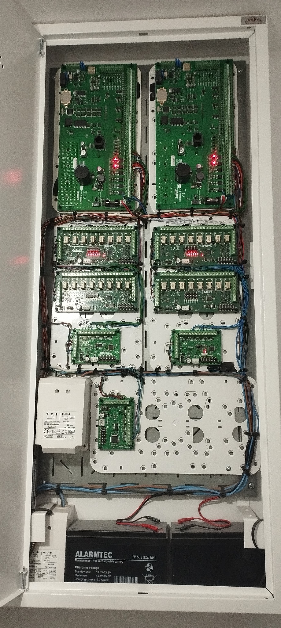 Rozdzielnica z modułami elektronicznymi systemu zabezpieczeń, widoczne liczne kable, płytki drukowane, transformator i akumulatory Alarmtec.