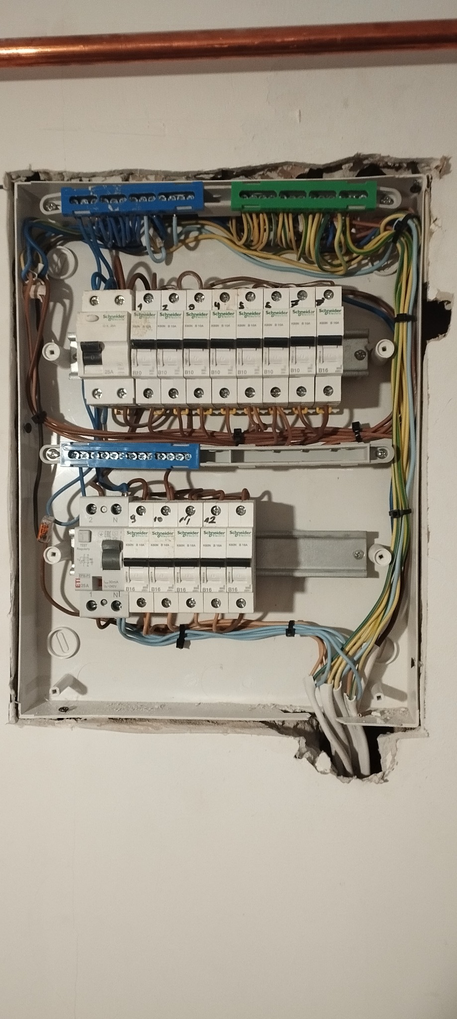 Otwarta skrzynka elektryczna z zamontowanymi bezpiecznikami Schneider Electric, widoczne kable w kolorowych izolacjach i uszkodzenia tynku wokół skrzynki.