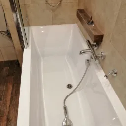 Biała wanna z chromowaną armaturą, prysznicem ręcznym i szklanym parawanem nawannowym na tle beżowych płytek i drewnianej półki z akcesoriami łazienkowymi.