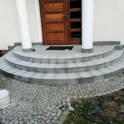 Wyłożone szarą kostką granitową, półokrągłe schody wejściowe z płytkami ceramicznymi, prowadzące do drewnianych drzwi z kolumnami, z widocznym wiaderkiem po farbie i kostką brukową na pierwszym...