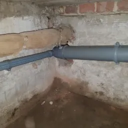 Fragment piwnicy z widocznymi szarymi rurami kanalizacyjnymi z tworzywa sztucznego, biegnącymi wzdłuż ściany z cegieł i popękanego tynku. Jedna rura owinięta materiałem izolacyjnym.