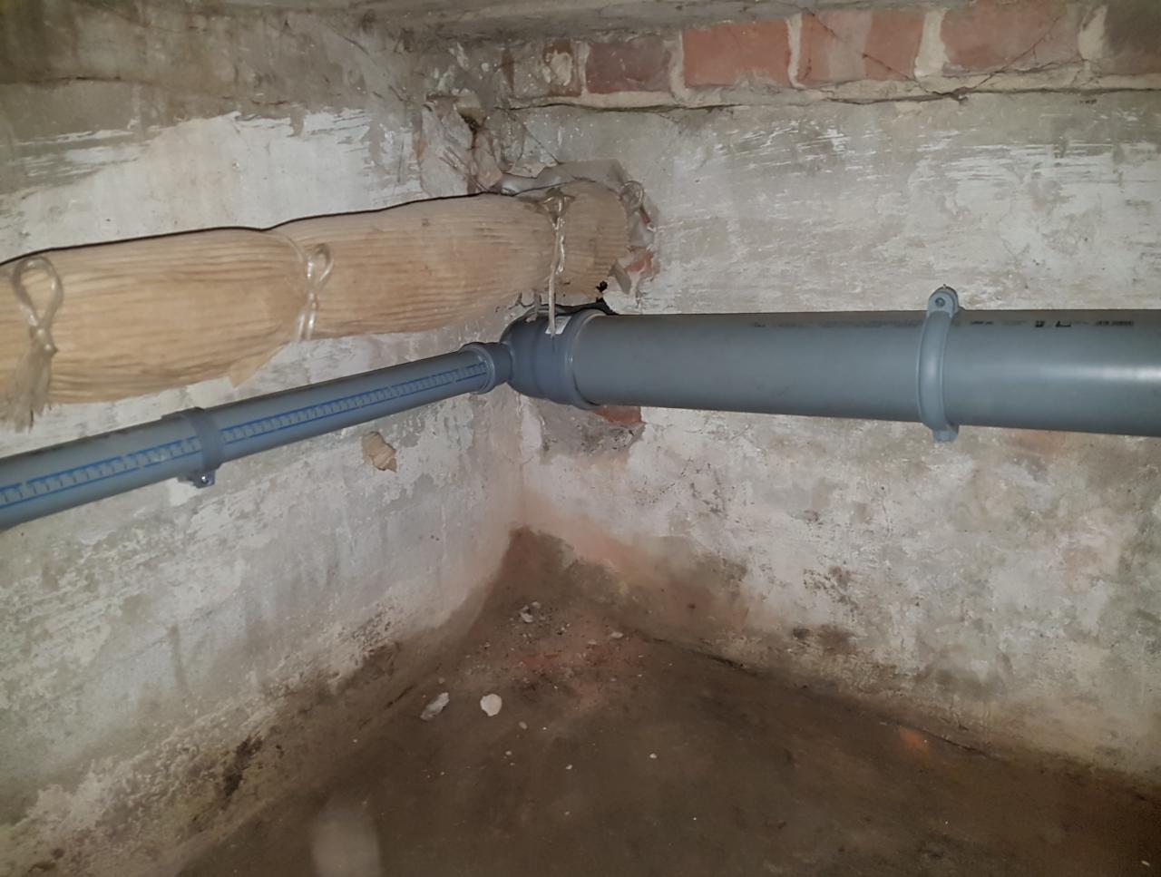 Fragment piwnicy z widocznymi szarymi rurami kanalizacyjnymi z tworzywa sztucznego, biegnącymi wzdłuż ściany z cegieł i popękanego tynku. Jedna rura owinięta materiałem izolacyjnym.