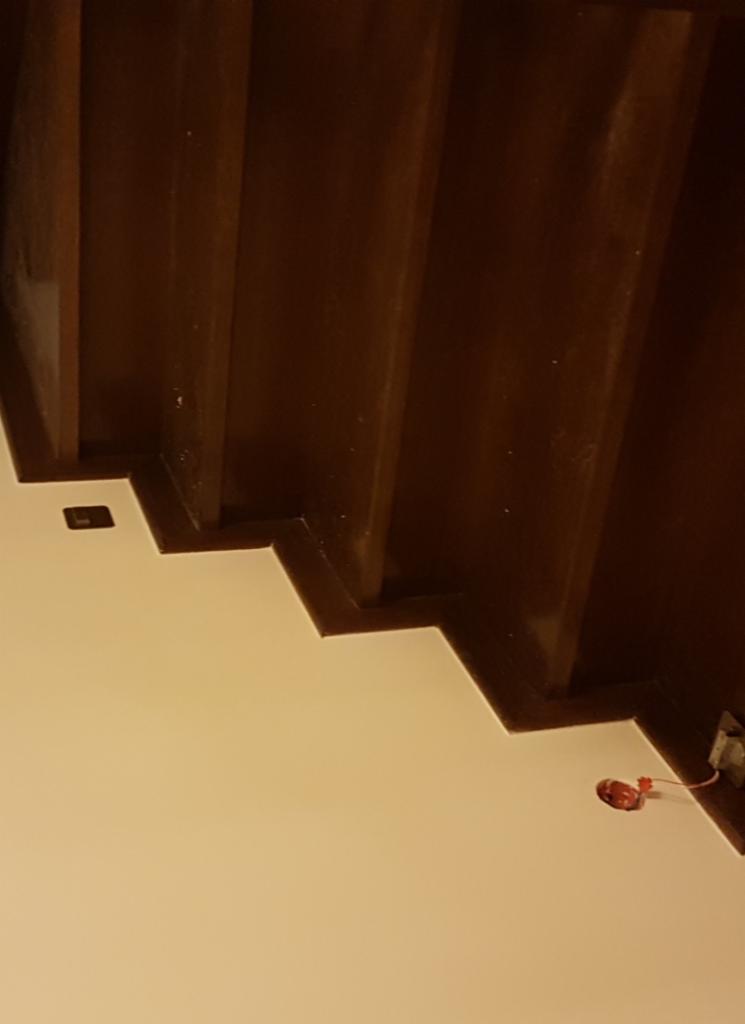 Pomieszczenie w trakcie wykończenia: świeżo pomalowana ściana w odcieniach bieli kontrastuje z ciemnobrązową, kaskadową konstrukcją sufitową, widoczne niedokończone gniazdko elektryczne...