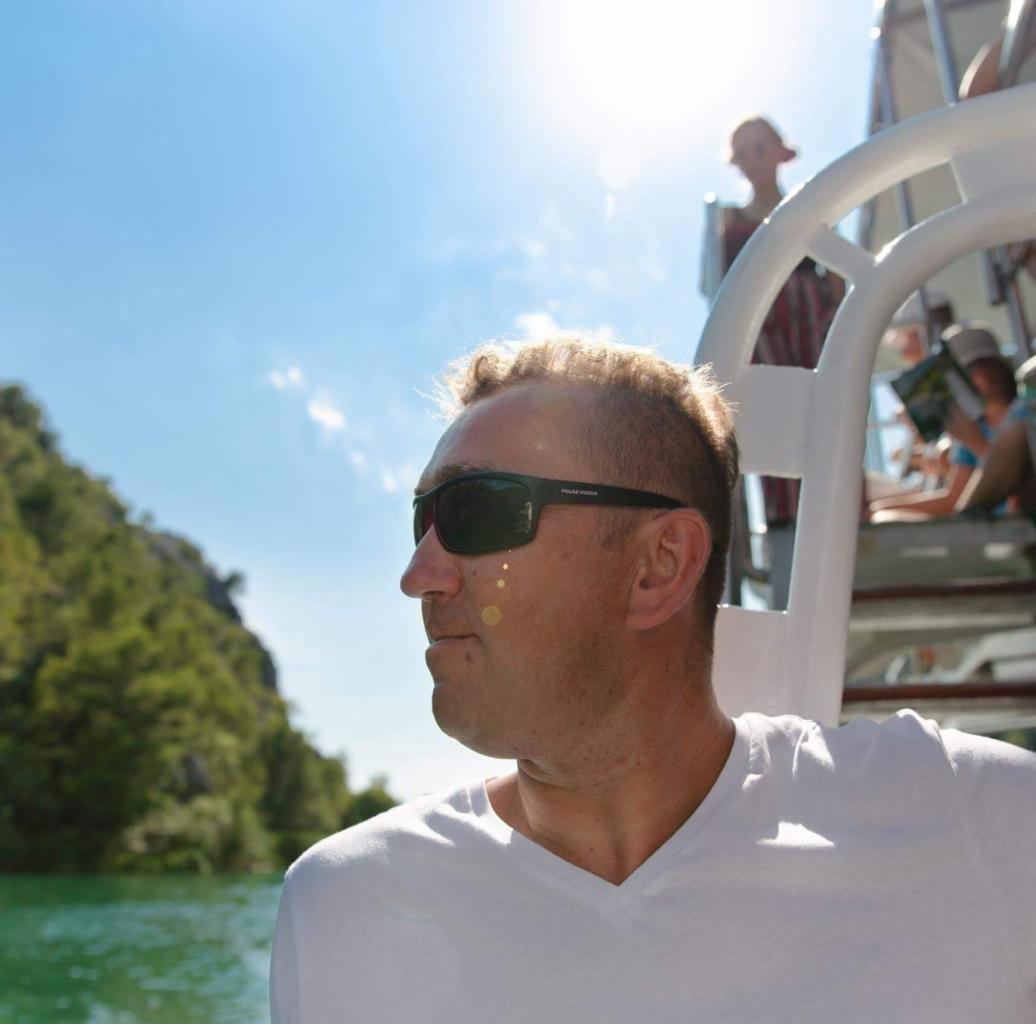 Mężczyzna w okularach przeciwsłonecznych na pokładzie łodzi, z widokiem na zielone wzgórza i błękitne niebo w słoneczny dzień. W tle widać innych pasażerów.
