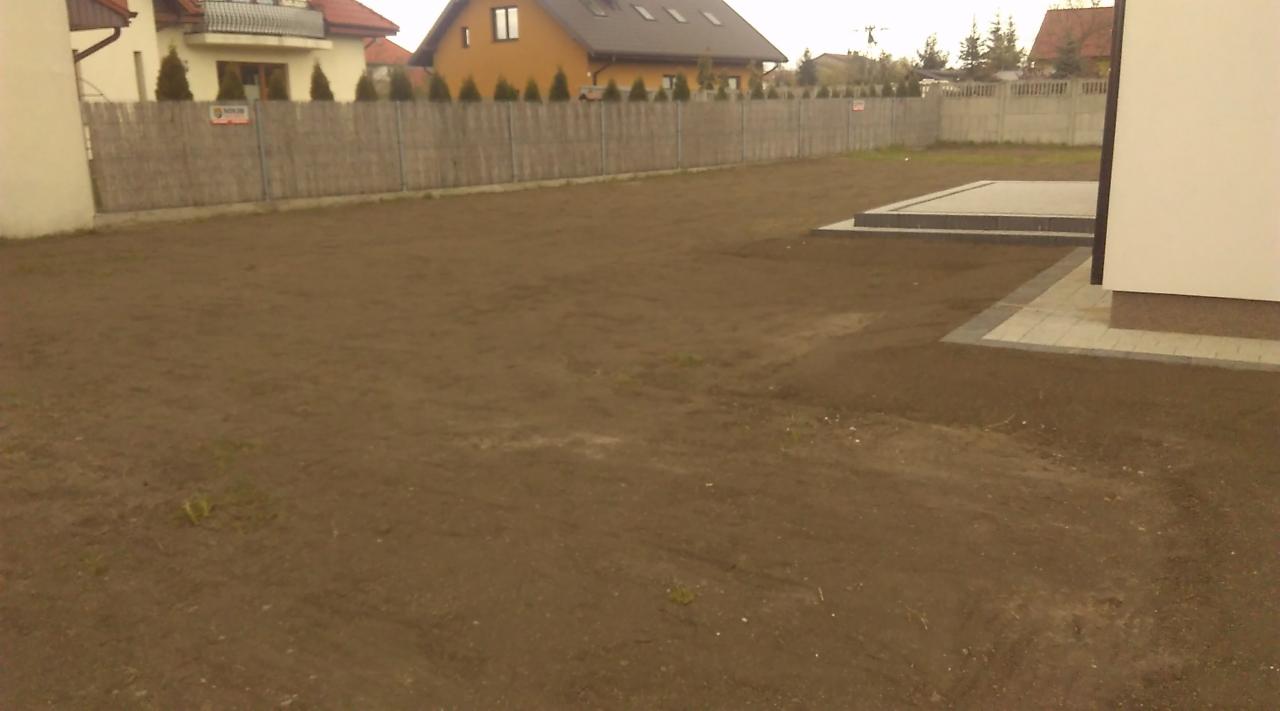 Przygotowany teren pod trawnik z widocznym fragmentem domu i ogrodzeniem w tle.