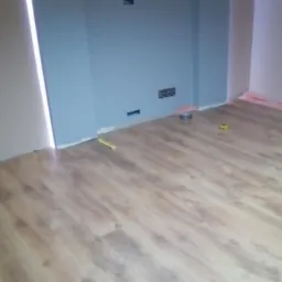 Układanie paneli podłogowych z widocznym podkładem izolacyjnym przy oknie i niebieską ścianą z gniazdkami elektrycznymi, żółta miarka leży na podłodze.