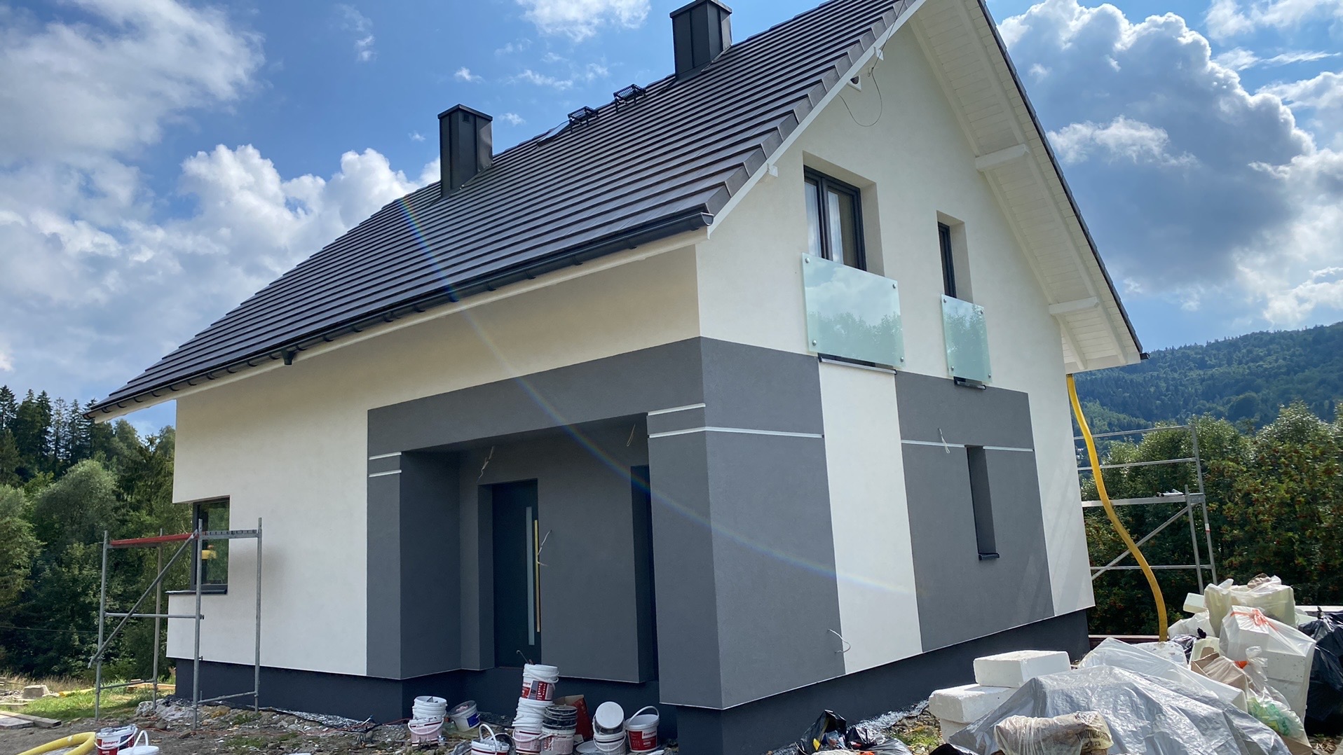 Nowoczesny dom jednorodzinny z ocieploną elewacją w odcieniach bieli i szarości, z ciemną dachówką, widoczne rusztowanie i pozostałości materiałów budowlanych na trawniku.