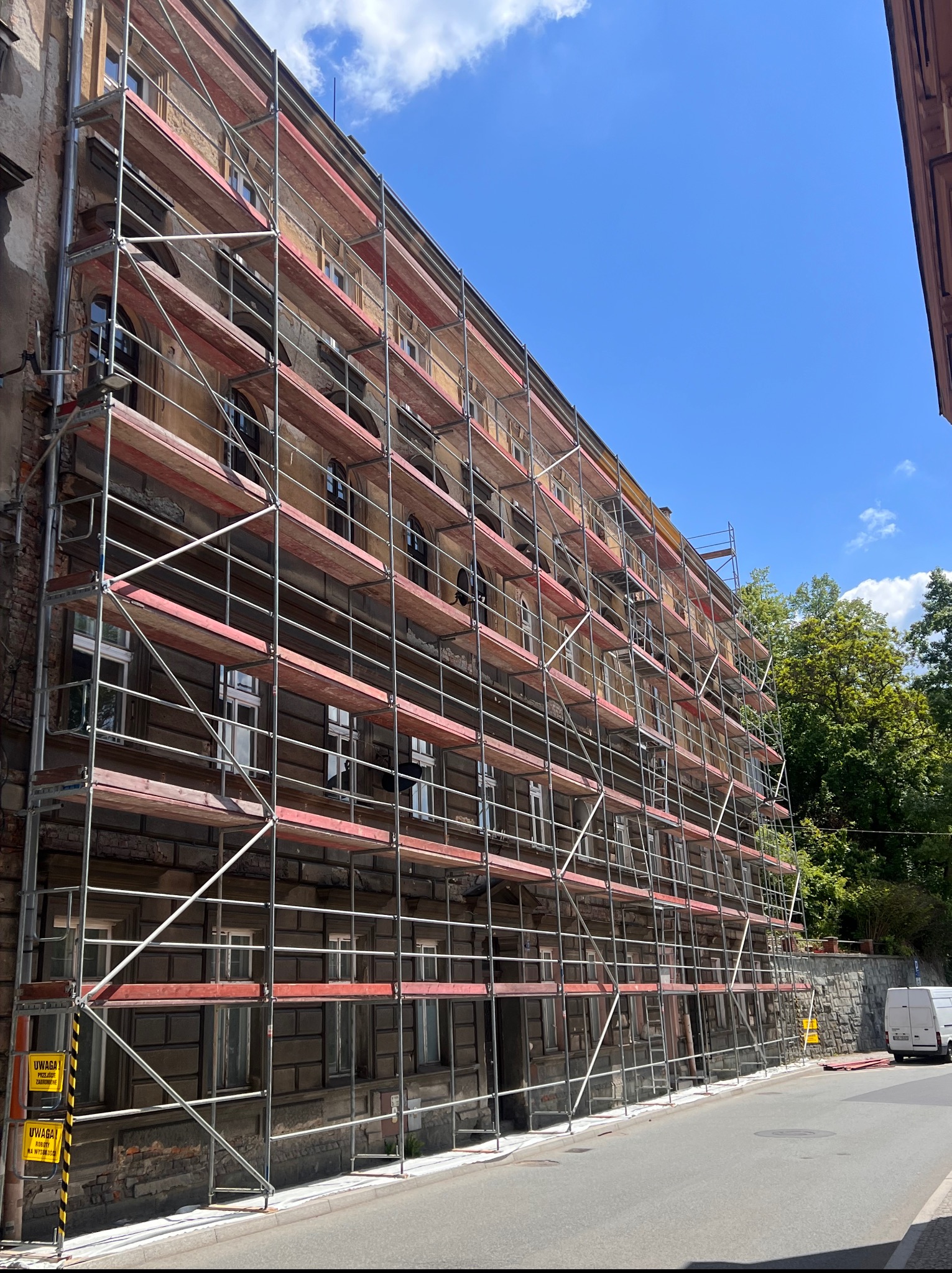 Budynek w rusztowaniach, widok z dołu na elewację w trakcie prac. Metalowe rusztowania z drewnianymi pomostami na tle błękitnego nieba.