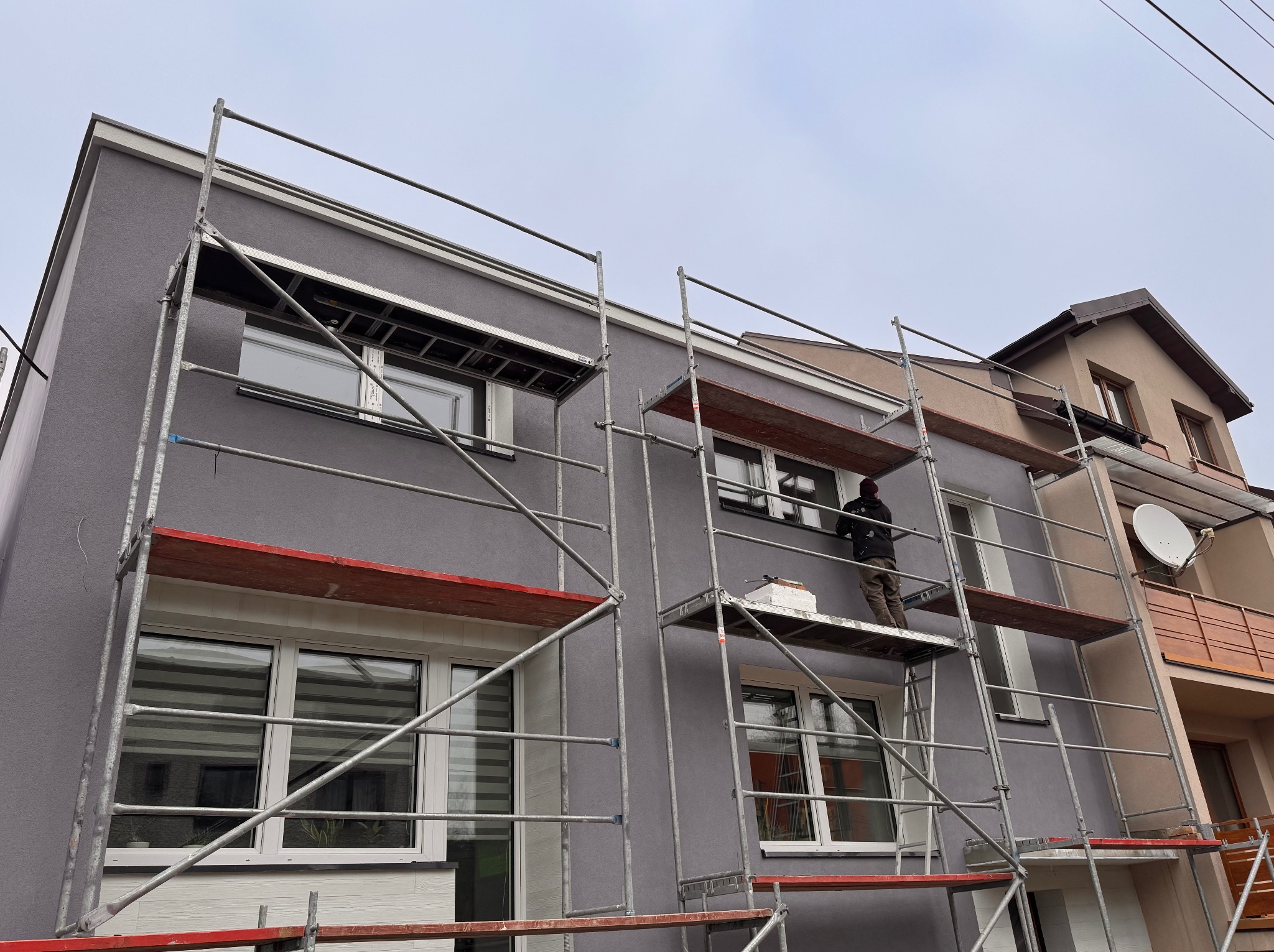 Szarego koloru elewacja domu w trakcie prac wykończeniowych. Na rusztowaniu stoi pracownik. Widoczne okna z roletami i fragment sąsiedniego budynku.