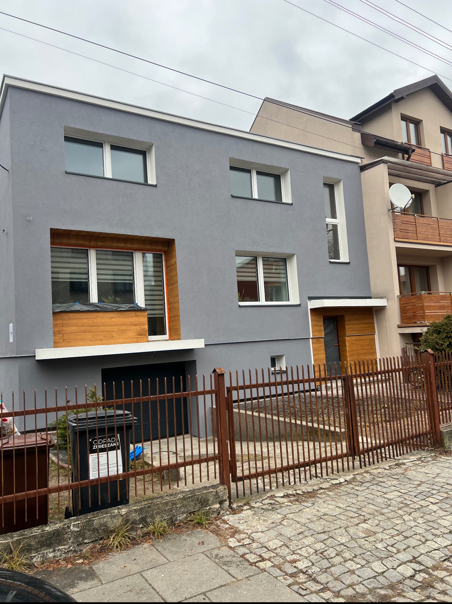 Szary, dwukondygnacyjny dom z drewnianymi elementami elewacji i czarną bramą garażową, widziany zza metalowego ogrodzenia. Dom sąsiaduje z innym budynkiem.