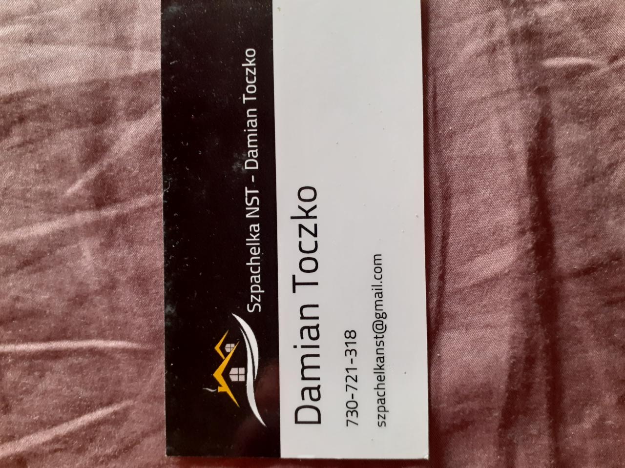 Wizytówka firmy 'Szpachelka NST - Damian Toczko' z numerem telefonu i adresem e-mail, na tle brązowej tkaniny.