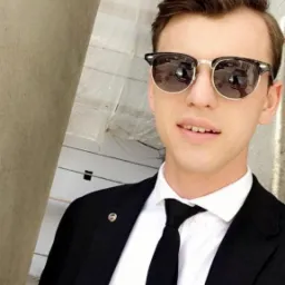 Młody mężczyzna w garniturze i okularach przeciwsłonecznych, robiący selfie przy jasnej ścianie.