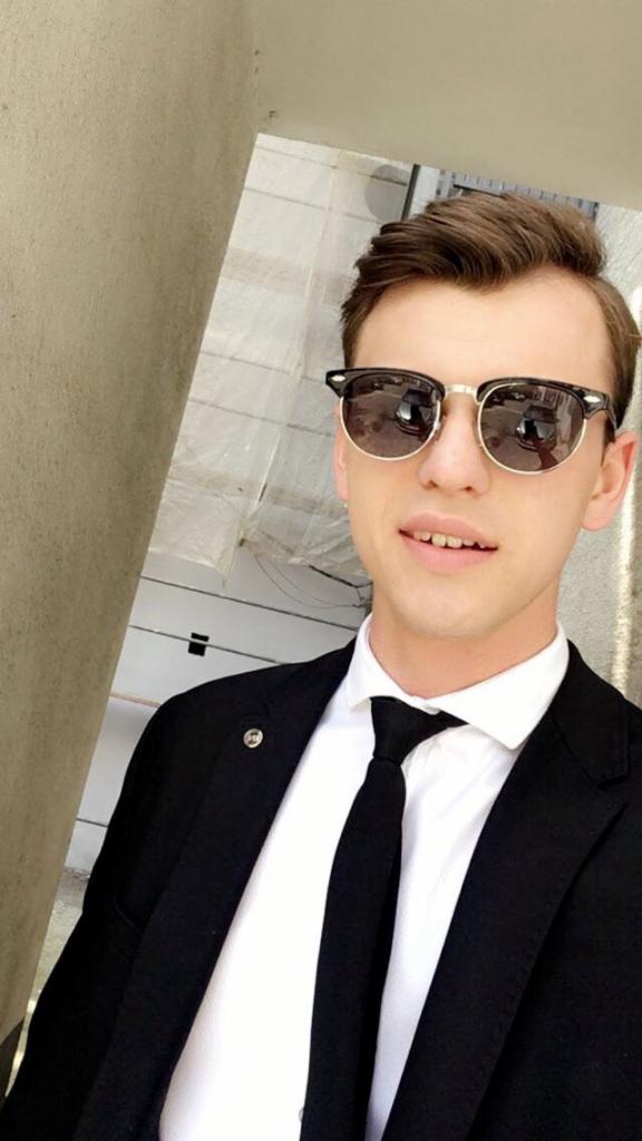 Młody mężczyzna w garniturze i okularach przeciwsłonecznych, robiący selfie przy jasnej ścianie.