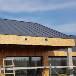 Nowoczesny dom w trakcie budowy z drewnianą elewacją, szarą blachą na dachu i widocznymi otworami wentylacyjnymi pod okapem, rusztowania budowlane na pierwszym planie.