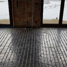 Rozłożona instalacja ogrzewania podłogowego na folii izolacyjnej, widok z góry, surowe ściany z cegły i okno z widokiem na zaśnieżony teren.