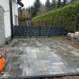 Świeżo ułożona, mokra kostka brukowa w odcieniach szarości na posesji, z widoczną zagęszczarką w rogu i stosem kostki przygotowanej do dalszych prac, tło stanowi ogrodzenie i zielony żywopłot.