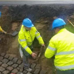 Trzech pracowników w odblaskowych kurtkach i niebieskich kaskach układa kostkę brukową w wykopie przy drodze, jeden z nich używa łopaty.