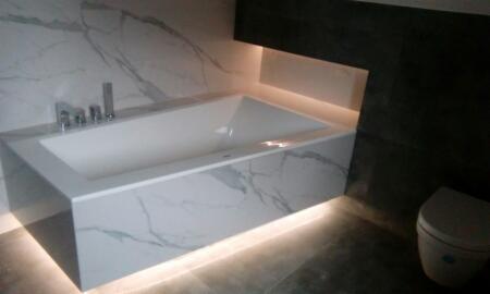 Nowoczesna wanna obudowana płytami imitującymi marmur, podświetlona od dołu ledami, z baterią wannową na tle ściany z motywem marmuru, obok toaleta i ciemna ściana.