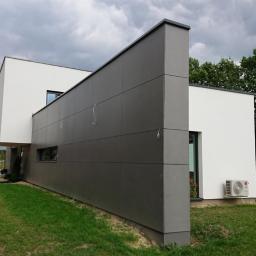 Innowacyjne Okna - Nowoczesny dom jednorodzinny z szarą elewacją panelową i białymi ścianami, widok z boku na trawnik i ogrodzenie, pochmurne niebo w tle.