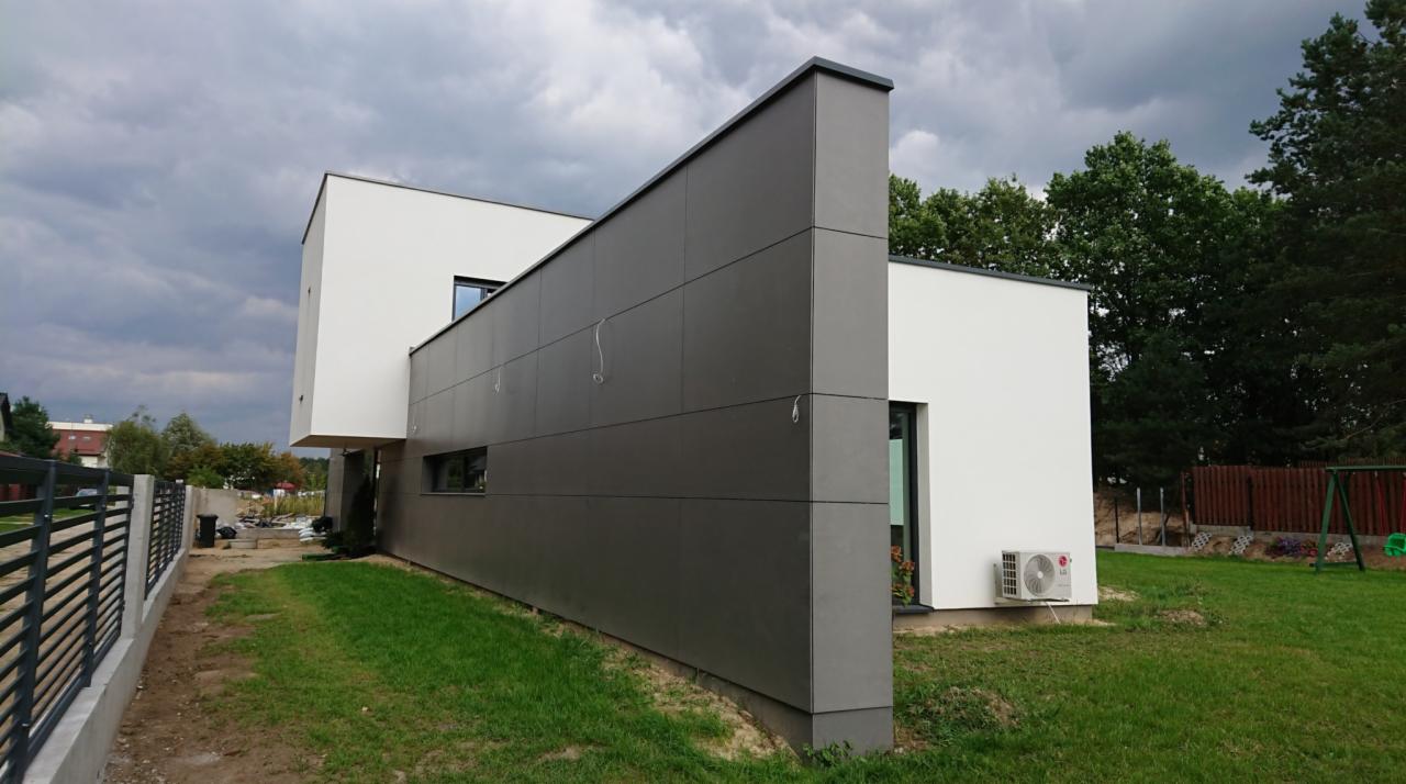 Nowoczesny dom jednorodzinny z szarą elewacją panelową i białymi ścianami, widok z boku na trawnik i ogrodzenie, pochmurne niebo w tle.