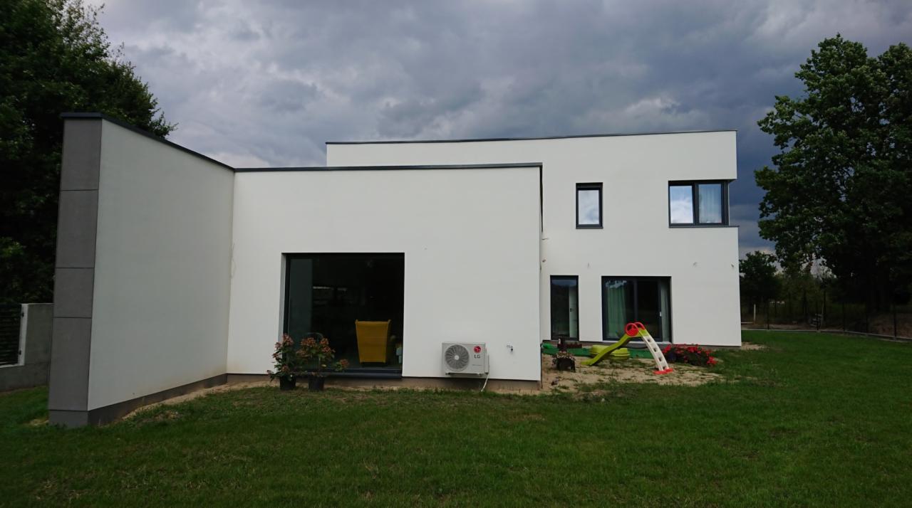Nowoczesny dom z przesuwnymi drzwiami tarasowymi, klimatyzatorem na elewacji, widok z ogrodu, pochmurne niebo.