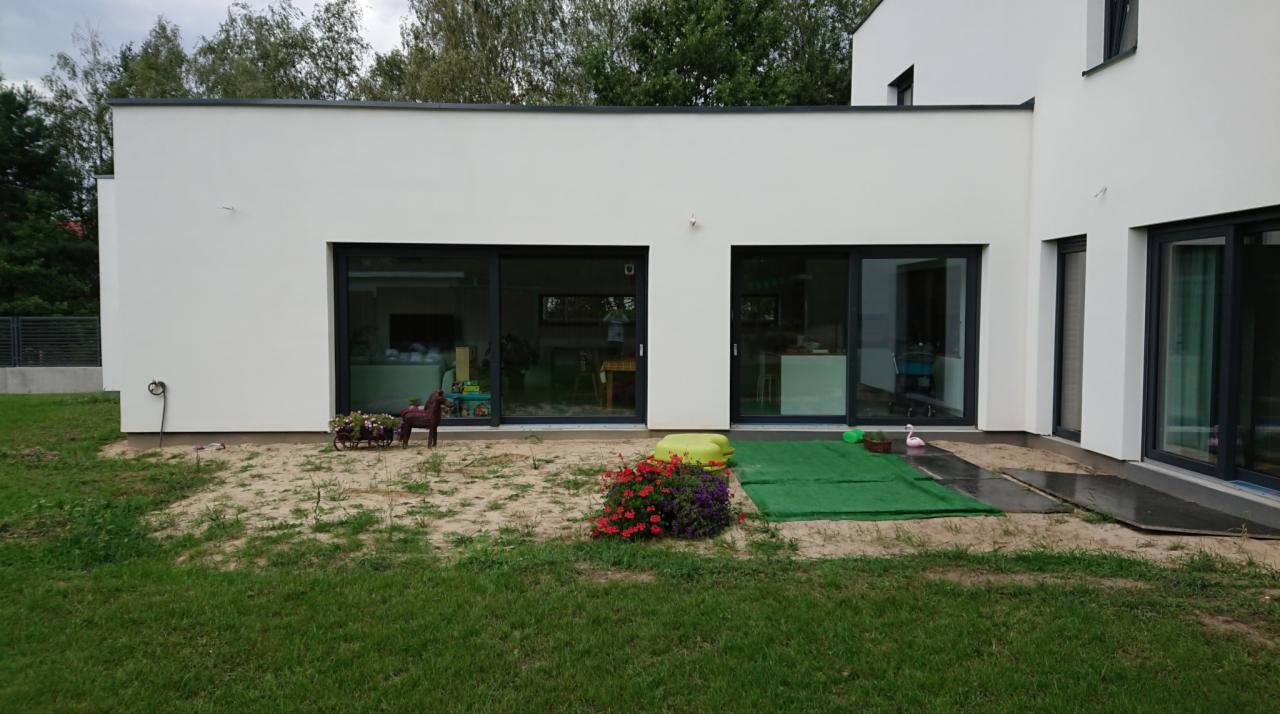 Nowoczesny dom z białymi ścianami i dużymi, czarnymi drzwiami przesuwnymi wychodzącymi na trawnik z elementami placu zabaw dla dzieci.