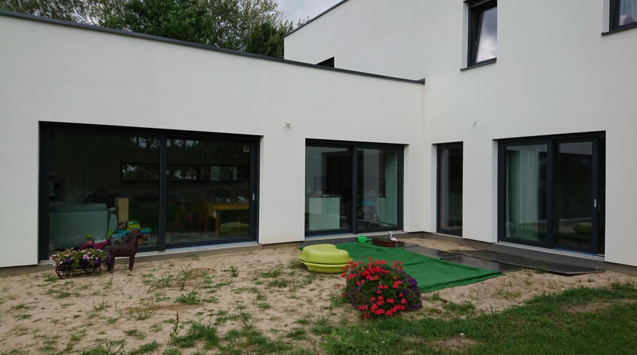 Nowoczesny dom z białymi ścianami i antracytowymi przesuwnymi oknami tarasowymi, widok z zewnątrz na piaszczysty plac zabaw z zabawkami i kwiatami.