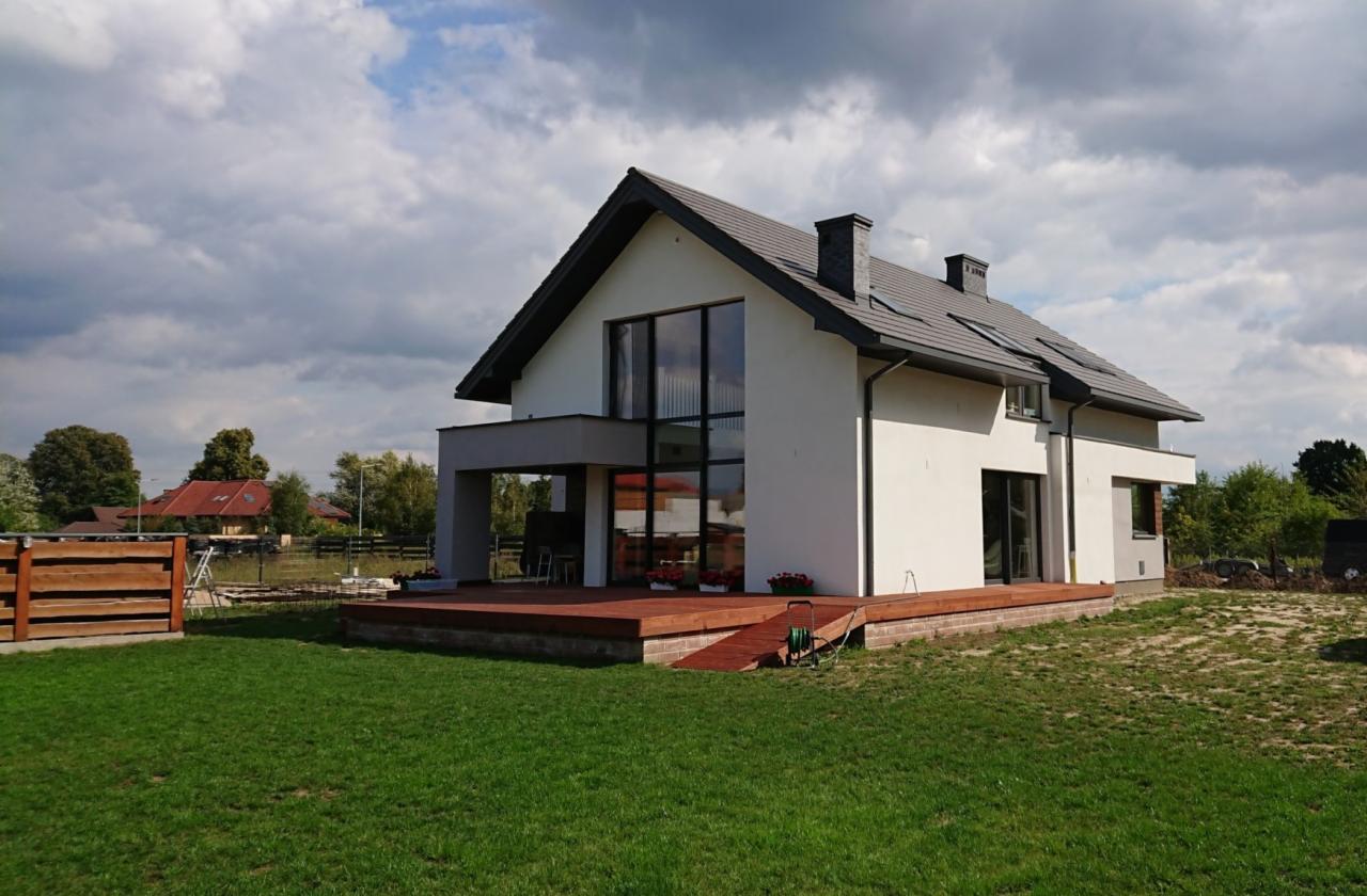 Nowoczesny dom jednorodzinny z dużymi oknami, tarasem z drewna i zadbanym trawnikiem na tle pochmurnego nieba.