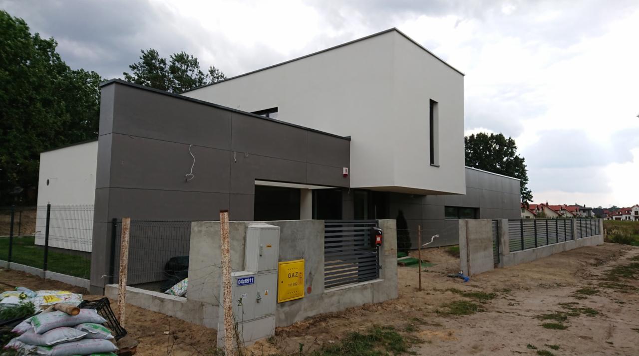 Nowoczesny dom jednorodzinny z szarą elewacją i minimalistycznymi oknami, ogrodzony metalowym płotem na betonowej podmurówce, widoczna skrzynka gazowa i worki z materiałami budowlanymi przed domem.