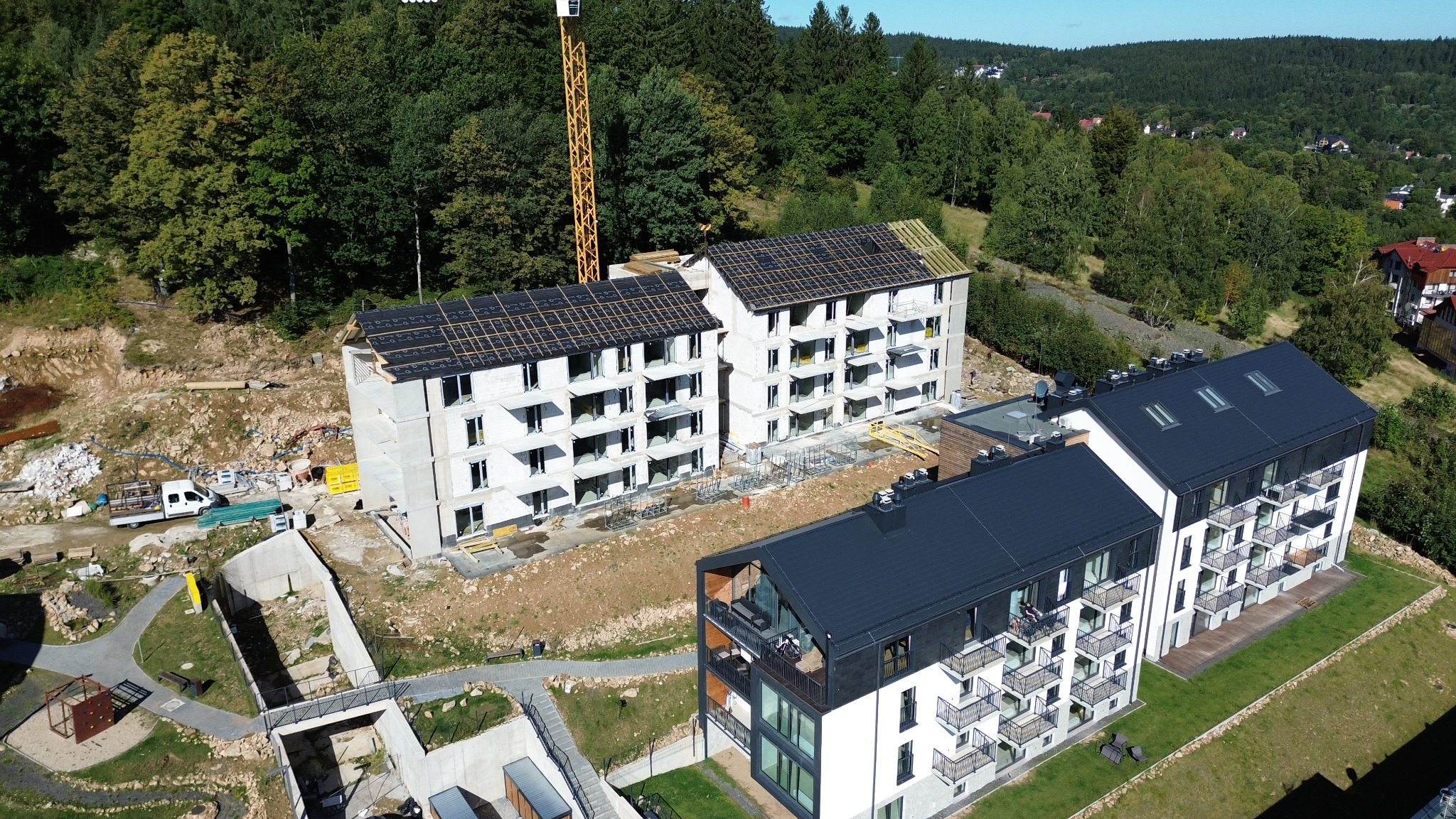 Widok z drona na budowę osiedla: dwa budynki w stanie surowym otwartym z widocznym deskowaniem dachu i jeden budynek wykończony z czarnym dachem i balkonami.