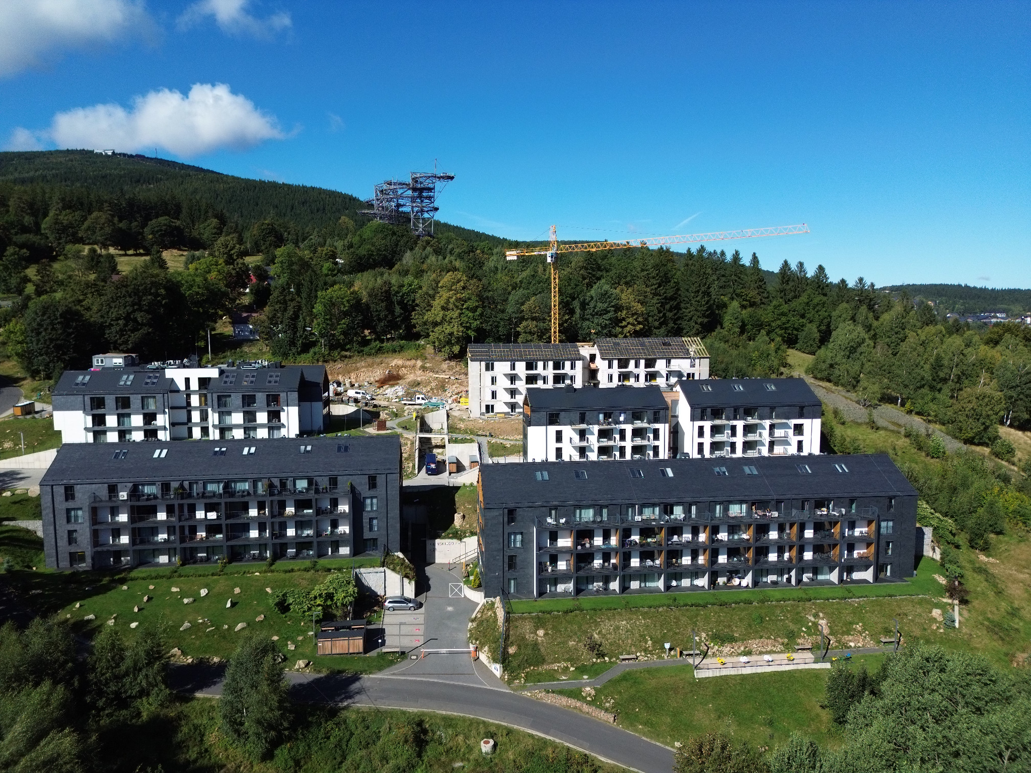 Widok z lotu ptaka na kompleks apartamentów w budowie we Wrocławiu, z żurawiem i otaczającą zielenią. Budynki z ciemnymi dachami i białymi elewacjami.