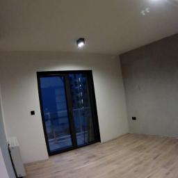 Janos - Wnętrze pokoju po remoncie: świeżo pomalowane ściany w odcieniach szarości i bieli, drewniana podłoga, czarne ramy okienne i drzwi balkonowe, widoczny fragment grzejnika.