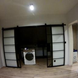 Janos - Nowoczesne, przesuwne drzwi w stylu loft z czarnymi ramami i mlecznym szkłem, dzielące przestrzeń z widoczną pralką w tle.