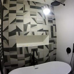 Janos - Nowoczesna łazienka z białą wanną wolnostojącą i czarną armaturą na tle ściany wyłożonej płytkami w geometryczne wzory, z wnęką na kosmetyki.